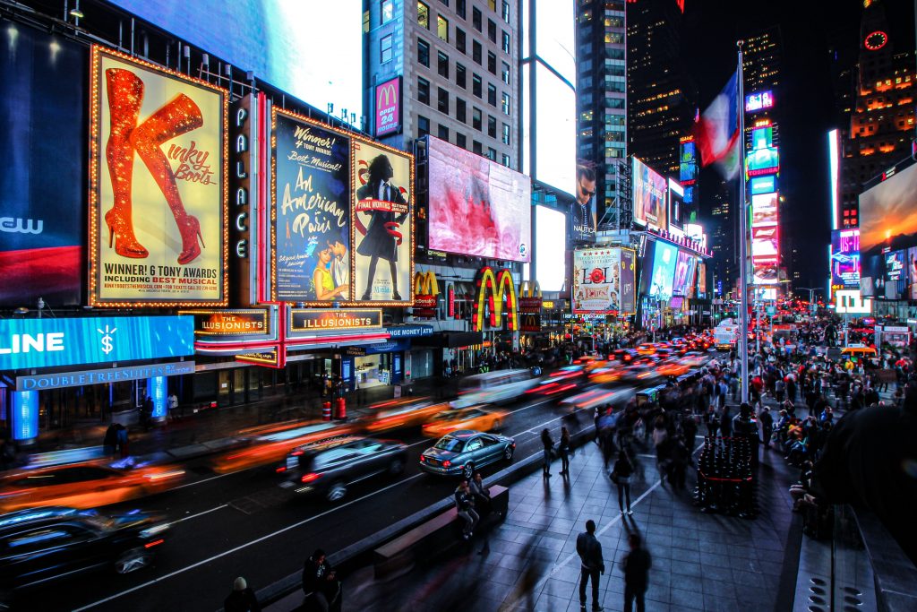 Fachadas iluminadas dos teatros da Broadway à noite, com pessoas andando na calçada.