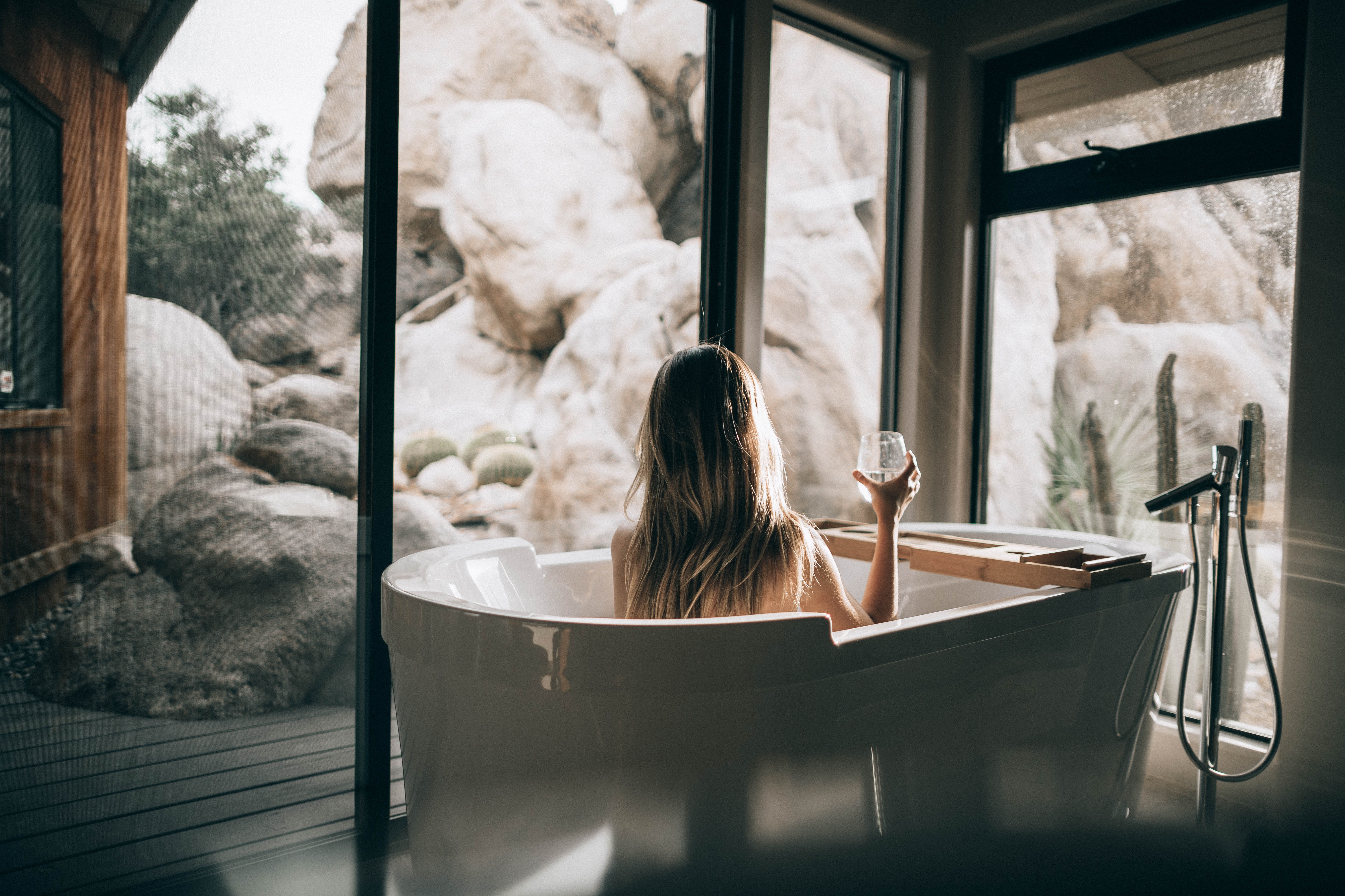 Relaxed woman in a bathtub with a glass of wine, gazing out of a window at a picturesque view. Solange Isaacs.