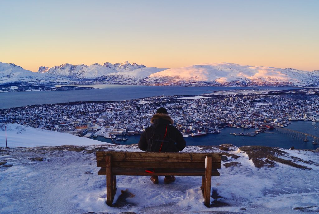 A person sitting alone on a bench surrounded by snow, gazing at the cityscape from atop a hill..
