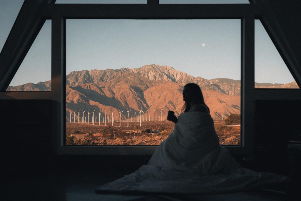 A girl holding a cup of coffe covered with a blanket, with a scenic moutian view outside of the window.