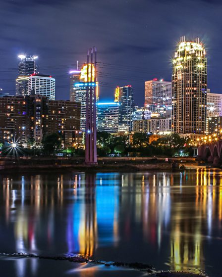 A nighttime view of Minneapolis with illuminated skyscrapers and their reflections in the river. By Solange Isaacs.