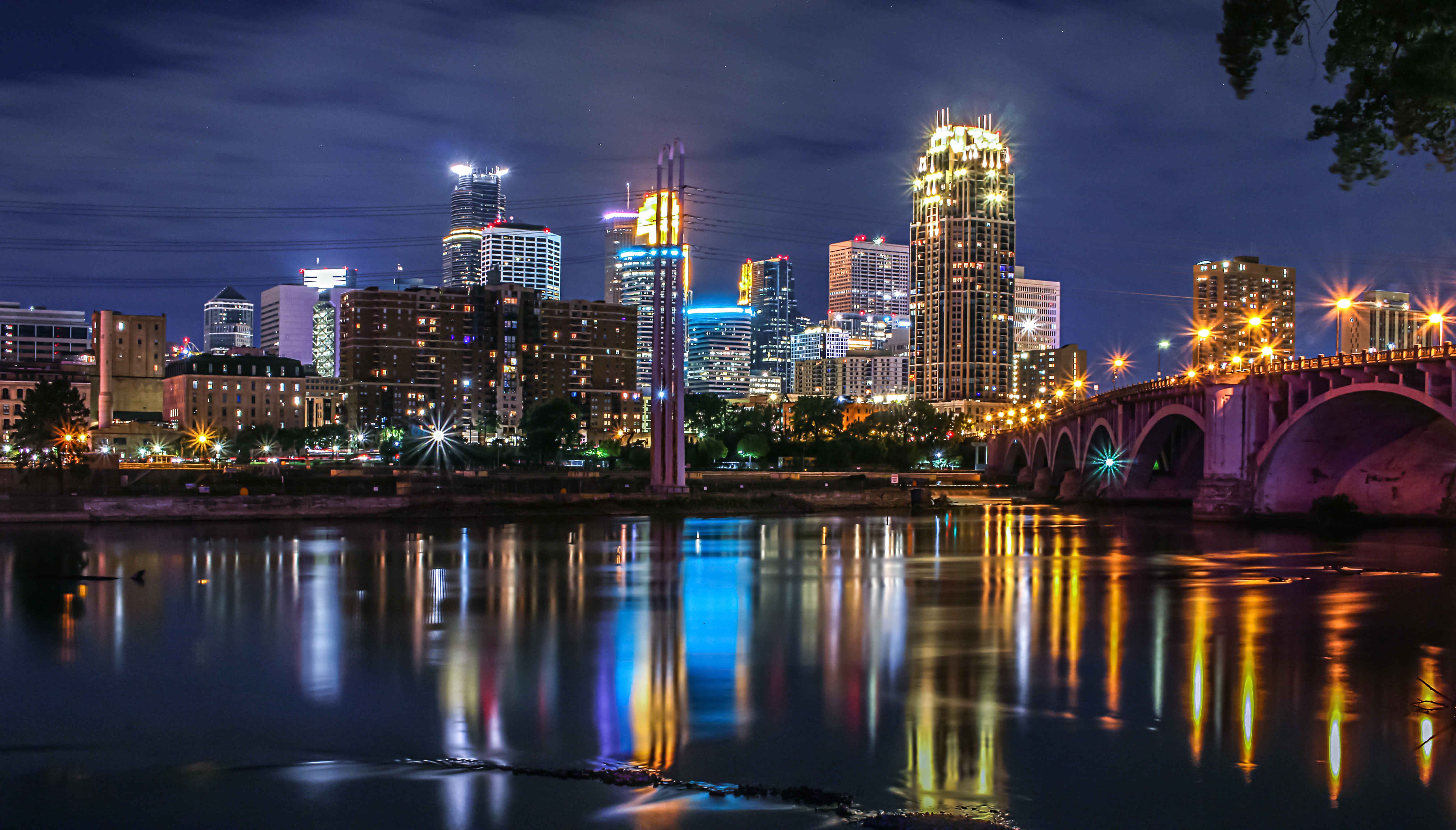 A nighttime view of Minneapolis with illuminated skyscrapers and their reflections in the river. By Solange Isaacs.